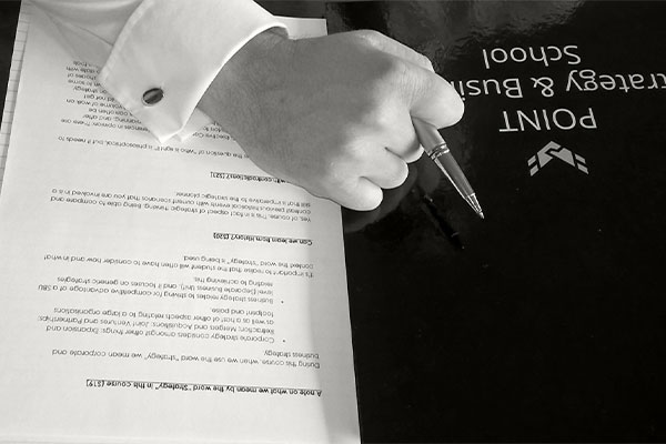 Mans hand holding pen resting on a POINT Strategy and Business School file and notes.