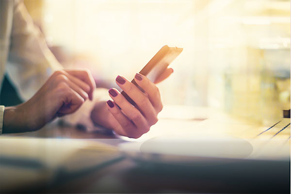 business woman's manicured hand holding a mobile phone at a work desk.